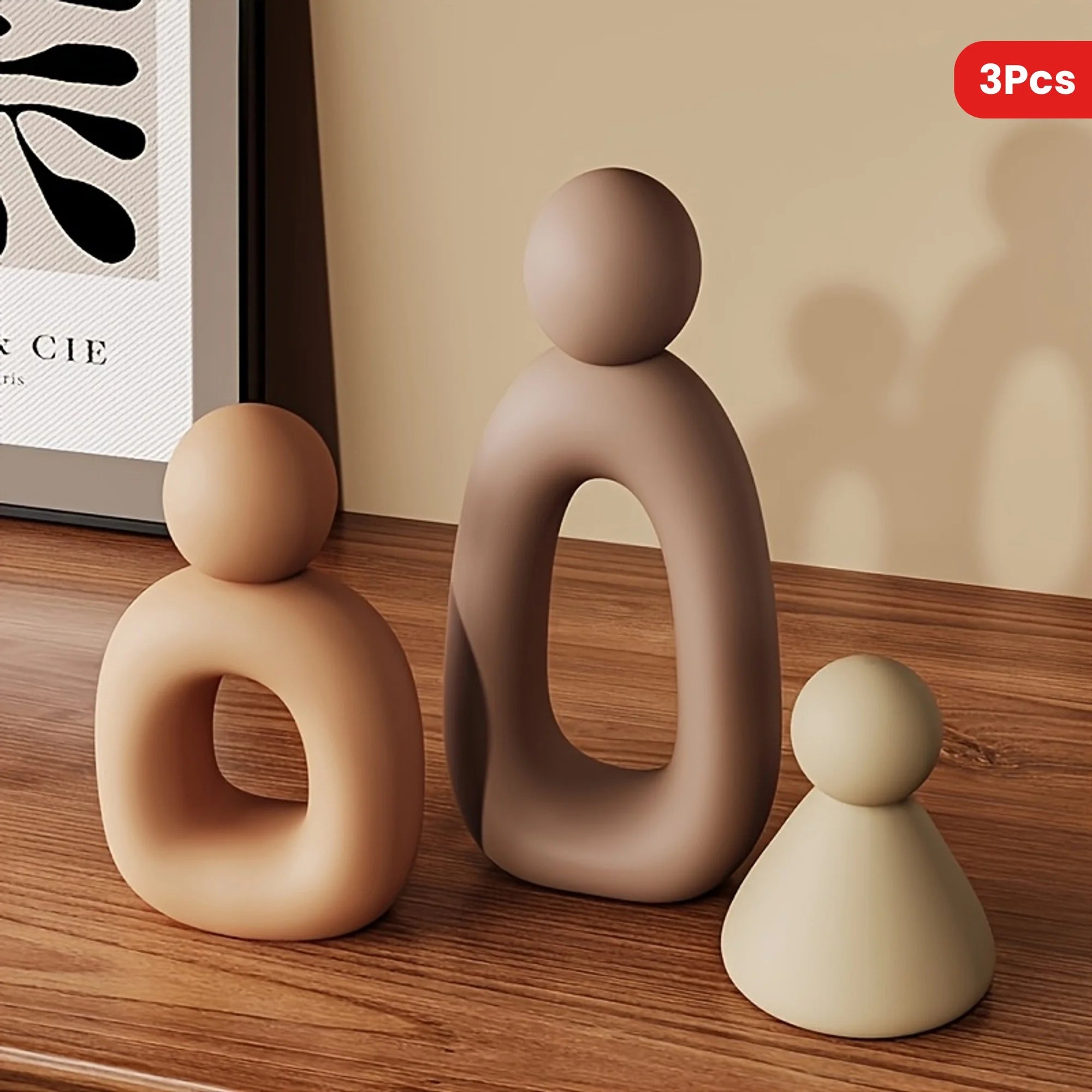 Three beige ceramic sculptures on a wooden surface with a framed picture in the background.