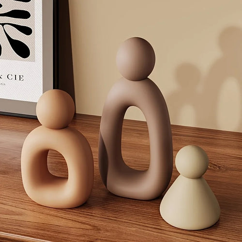 Three abstract ceramic figures on a wooden surface with a framed picture in the background.